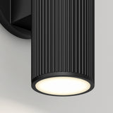 A close-up of a modern black cylindrical wall sconce with vertical grooves, mounted on a light gray wall, emitting a soft, warm white light from its circular base.