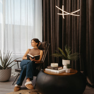 A woman relaxes in a modern chair by the window, reading beside potted plants and books, with a lit candle atop a round black table. Behind her hang dark curtains and an Artika Orly modern LED pendant light (5 CCT).