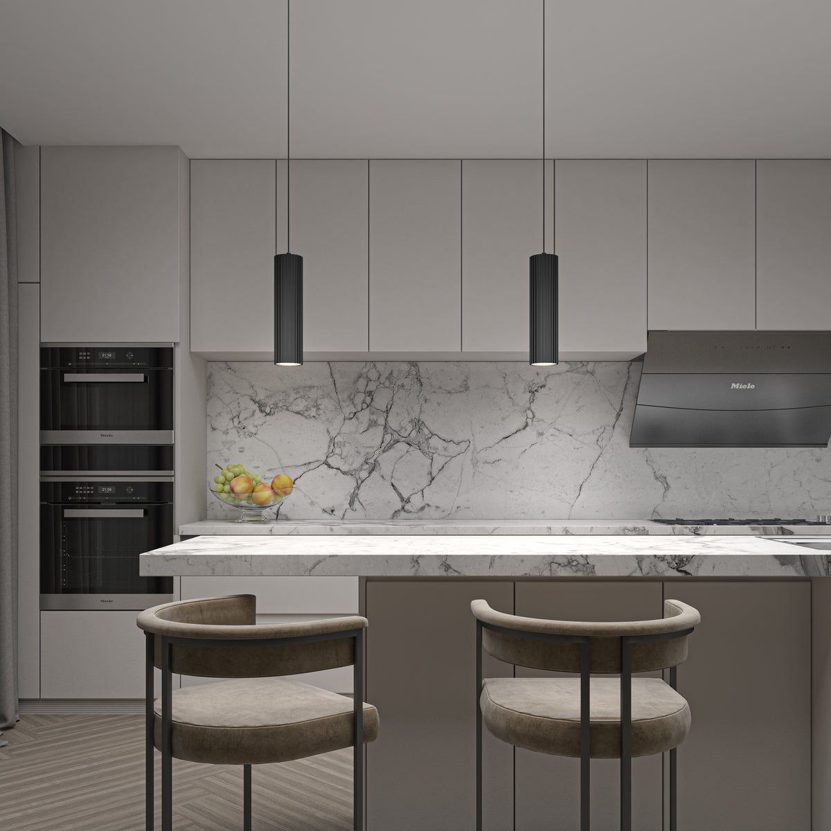 Minimalist modern kitchen with marble backsplash and countertop, two tan barstools at an island, black pendant lights, built-in ovens, a bowl of fruit, and sleek grey cabinets.