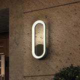 A modern oval-shaped outdoor wall light with a glowing border is mounted on a beige stone wall next to a wooden door and green shrubbery.