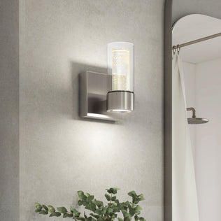 The Artika Essence 1-light LED bathroom vanity light features a cylindrical glass shade for accent lighting near a mirror and plant.