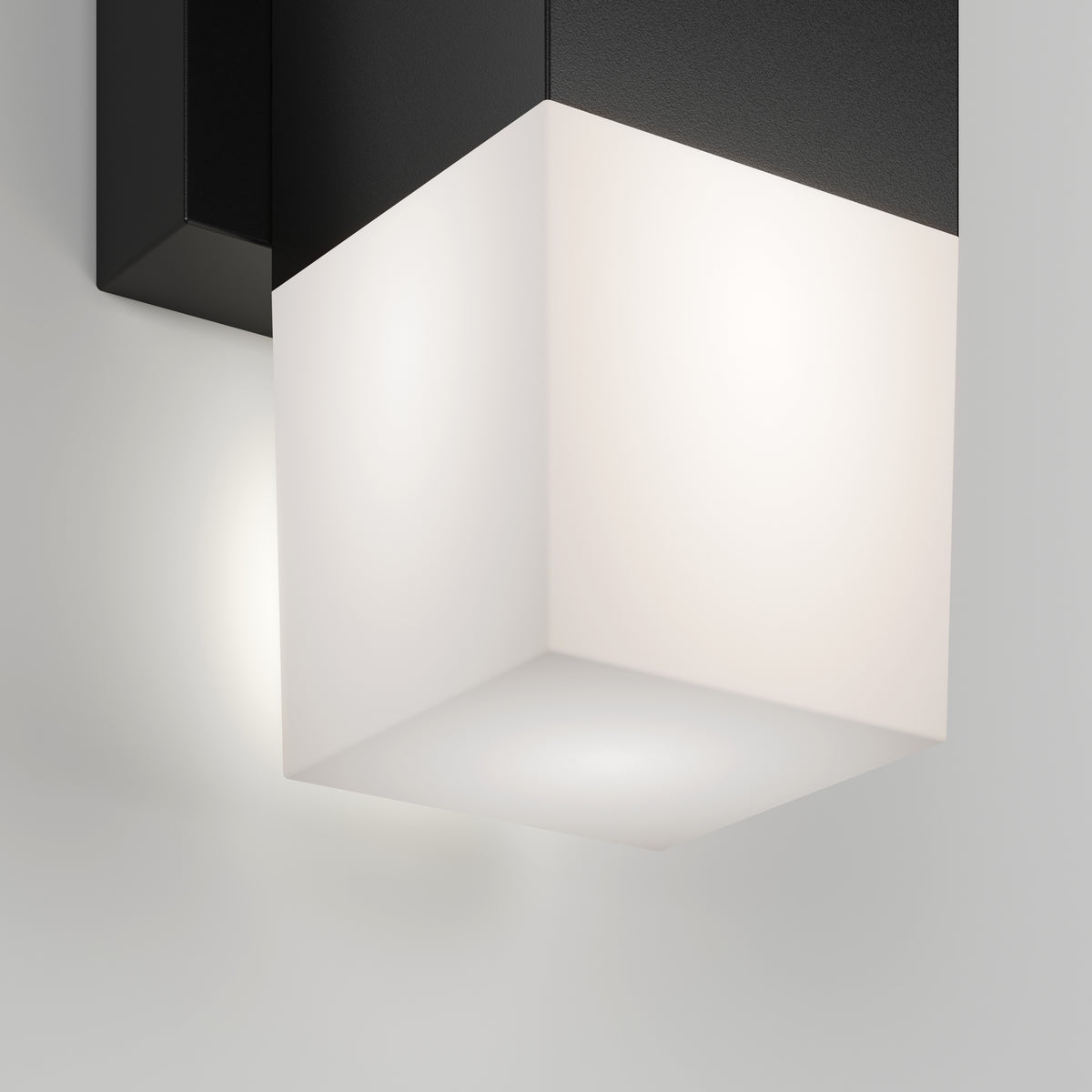 A modern wall-mounted light fixture with a black base and a glowing frosted white cube-shaped shade, casting soft light against a white wall.