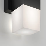 A modern wall-mounted light fixture with a black base and a glowing frosted white cube-shaped shade, casting soft light against a white wall.