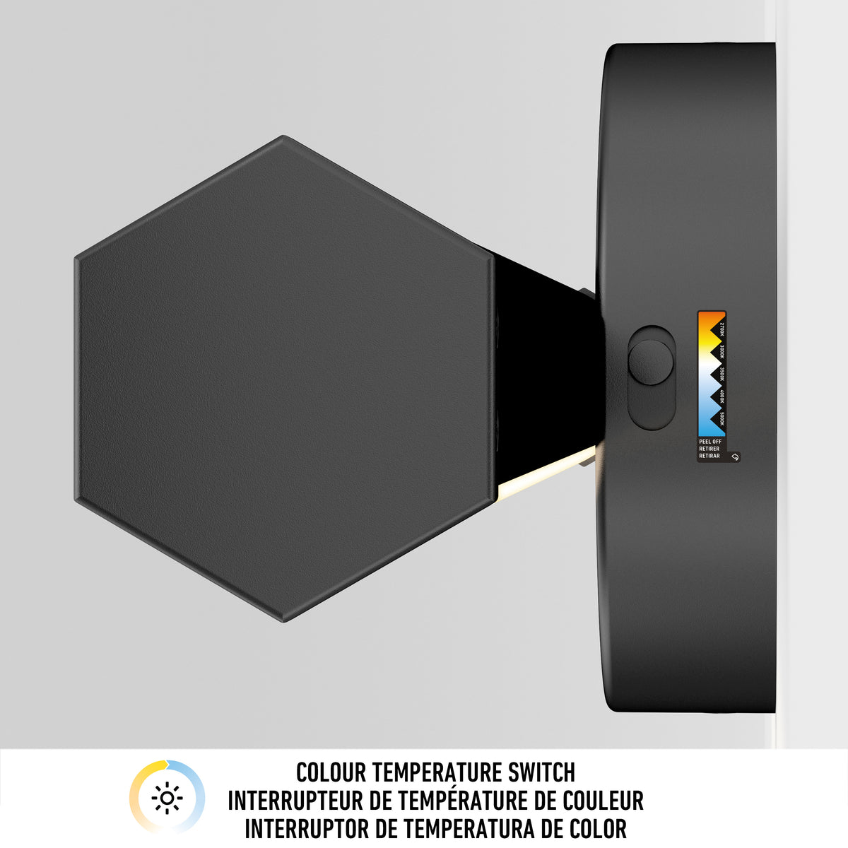 A modern hexagonal wall light with a colour temperature switch, mounted on a white wall. A label below shows icons and multilingual text explaining the colour temperature adjustment feature.