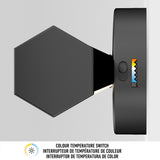 A modern hexagonal wall light with a colour temperature switch, mounted on a white wall. A label below shows icons and multilingual text explaining the colour temperature adjustment feature.