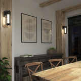 A rustic dining room with a wooden table, two wooden chairs, a sideboard with books and a plant basket, abstract art on the wall, and two modern wall sconces providing warm light.