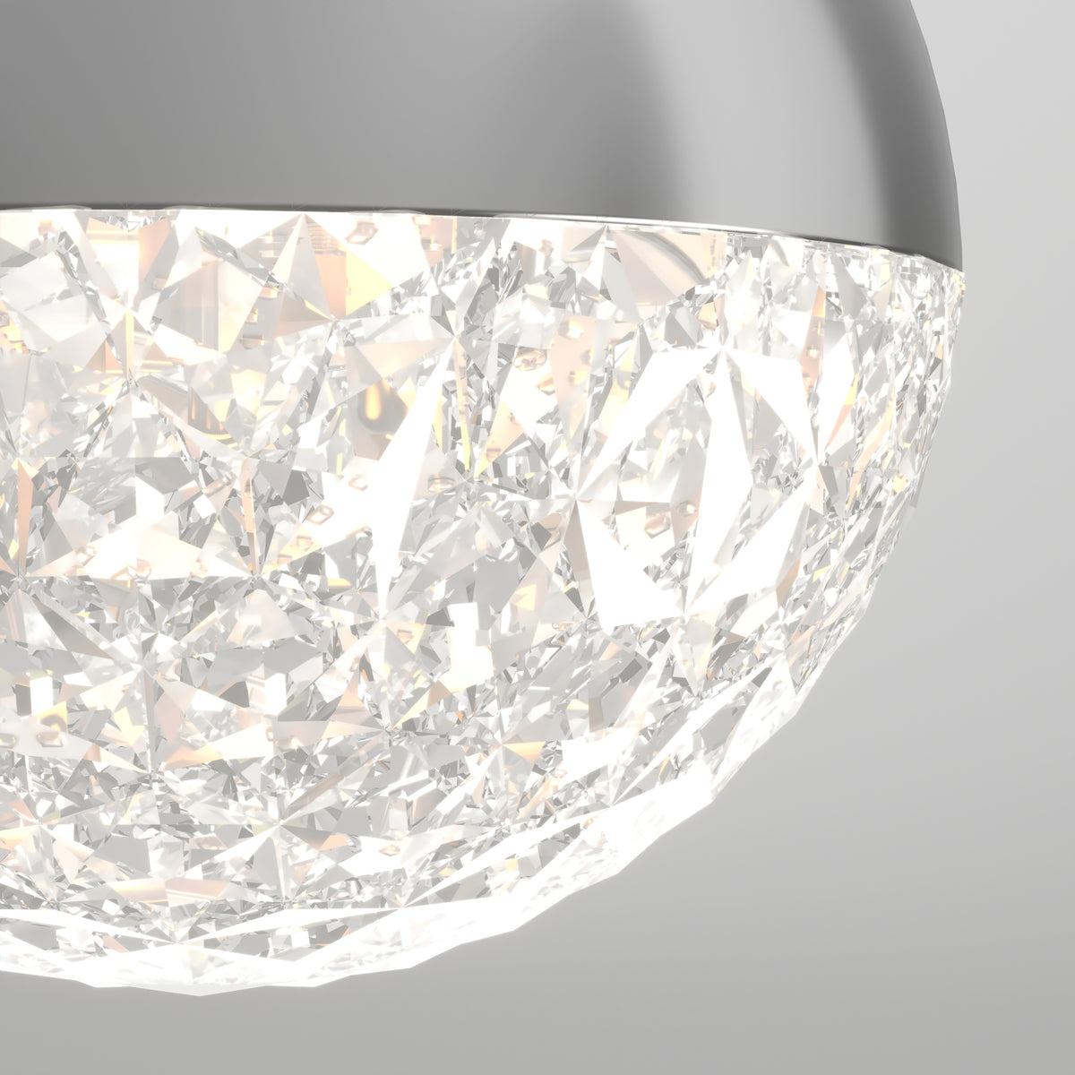 Close-up of a modern ceiling light fixture with a metallic rim and a faceted, crystal-like glass surface that reflects and diffuses light, creating a sparkling effect. The background is plain and light gray.