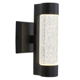 A modern black wall sconce with a cylindrical clear glass shade featuring bubble-like details, illuminated with warm white light, mounted on a square backplate.