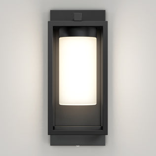 The Artika Aurel Outdoor LED Light, 3CCT features a rectangular black metal frame with a frosted cylindrical glass shade, mounted on a light wall and illuminated to cast a gentle glow.