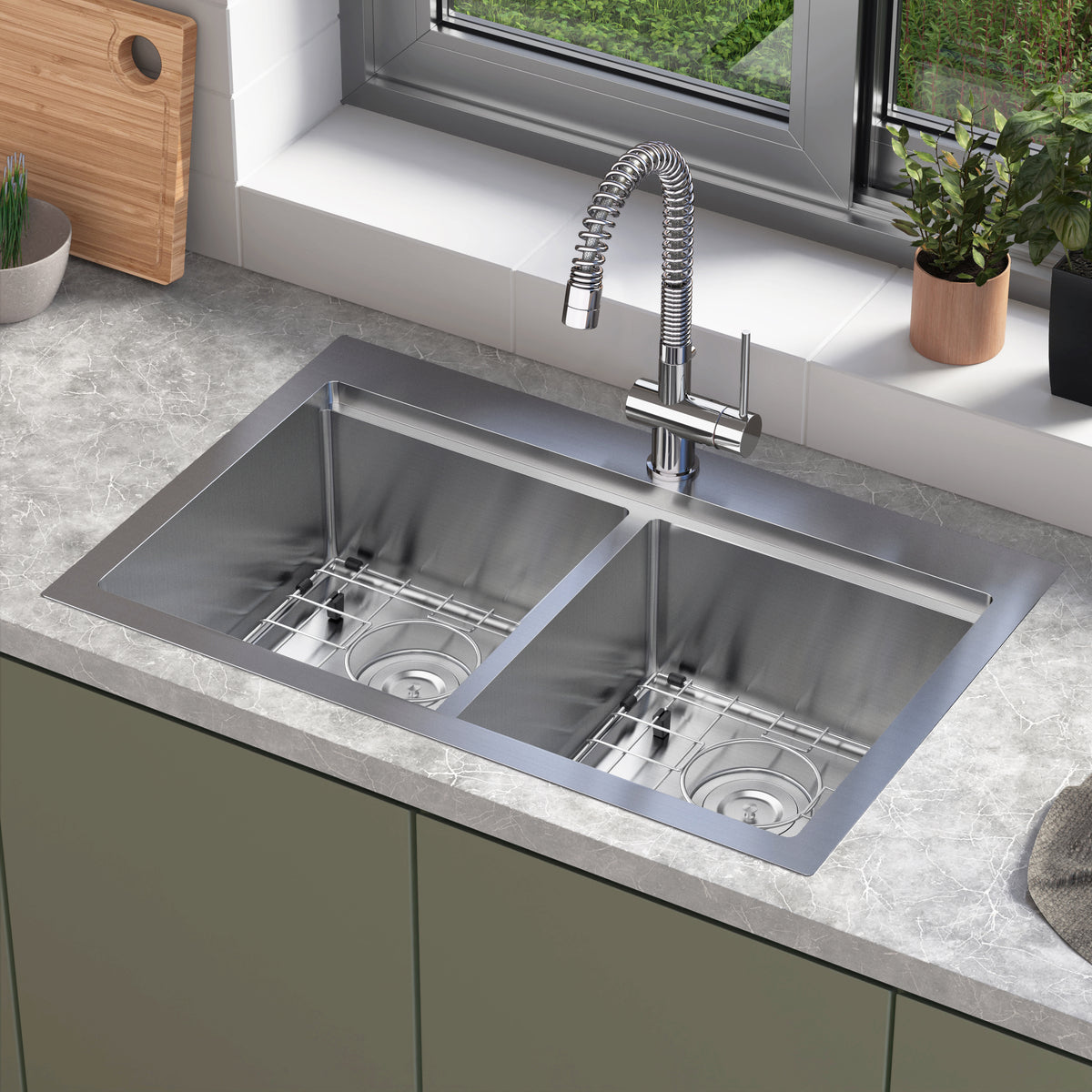 A modern kitchen sink with two stainless steel basins, a tall curved faucet, and a marble countertop. The sink is beneath a window with green plants and a cutting board on the windowsill.