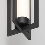 Close-up of a modern wall sconce featuring a black geometric metal frame surrounding a cylindrical frosted glass light, mounted on a light gray wall.