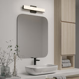 Minimalist bathroom with a large rectangular mirror, modern light fixture above, white vessel sink, black faucet, light wood cabinet, rolled towels, glass accessories, and sprigs in a vase on a white countertop.