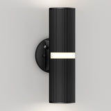 A modern black wall sconce with a ribbed cylindrical design, mounted on a round base, emits light from a central horizontal strip and both the top and bottom. The fixture is attached to a plain light-colored wall.