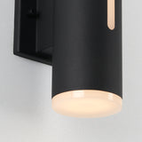 Close-up of a modern black cylindrical wall light fixture mounted on a light-colored wall, emitting a soft, warm glow from the frosted bottom and a slim vertical cutout.