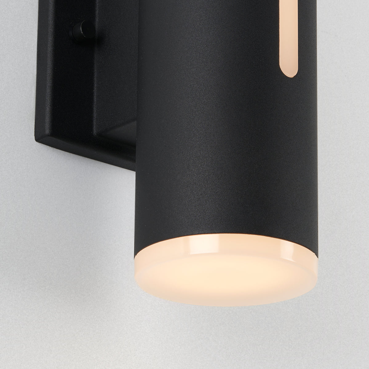 A close-up of the Artika Linea modern indoor-outdoor LED wall light, featuring a black cylindrical design with a frosted bottom diffuser, emits a soft, warm glow while mounted on a light-colored wall.