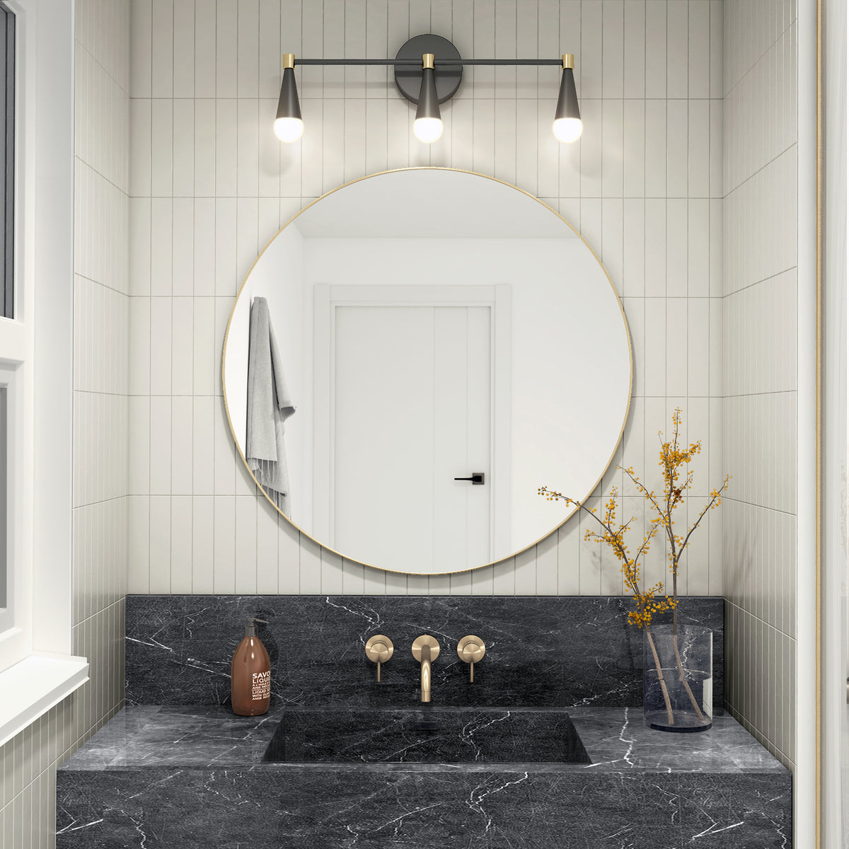 Modern bathroom with a round mirror above a black marble sink, gold fixtures, a brown soap dispenser, and a clear vase with yellow branches. Three lights are mounted above the mirror, and a white door is reflected.