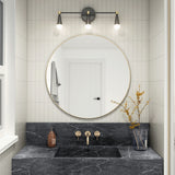 Modern bathroom with a round mirror above a black marble sink, gold fixtures, a brown soap dispenser, and a clear vase with yellow branches. Three lights are mounted above the mirror, and a white door is reflected.