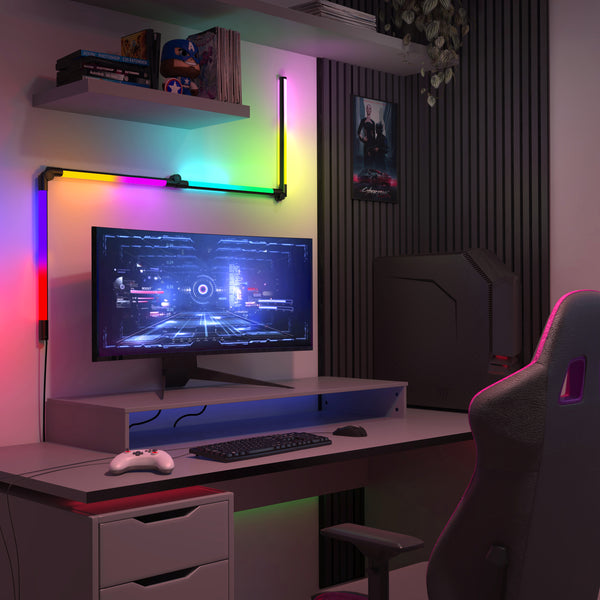 A modern gaming setup features an Artika Fitch smart foldable RGB LED gaming floor lamp beside a desk with a monitor displaying a futuristic game, white controller, keyboard, and mouse. A gaming chair and wall poster complete the scene.