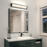 Modern bathroom with a square mirror above a white marble sink, green vanity, black faucet, glass jar, rolled towel, small plant, and a horizontal light fixture on the wall. A window lets in daylight.