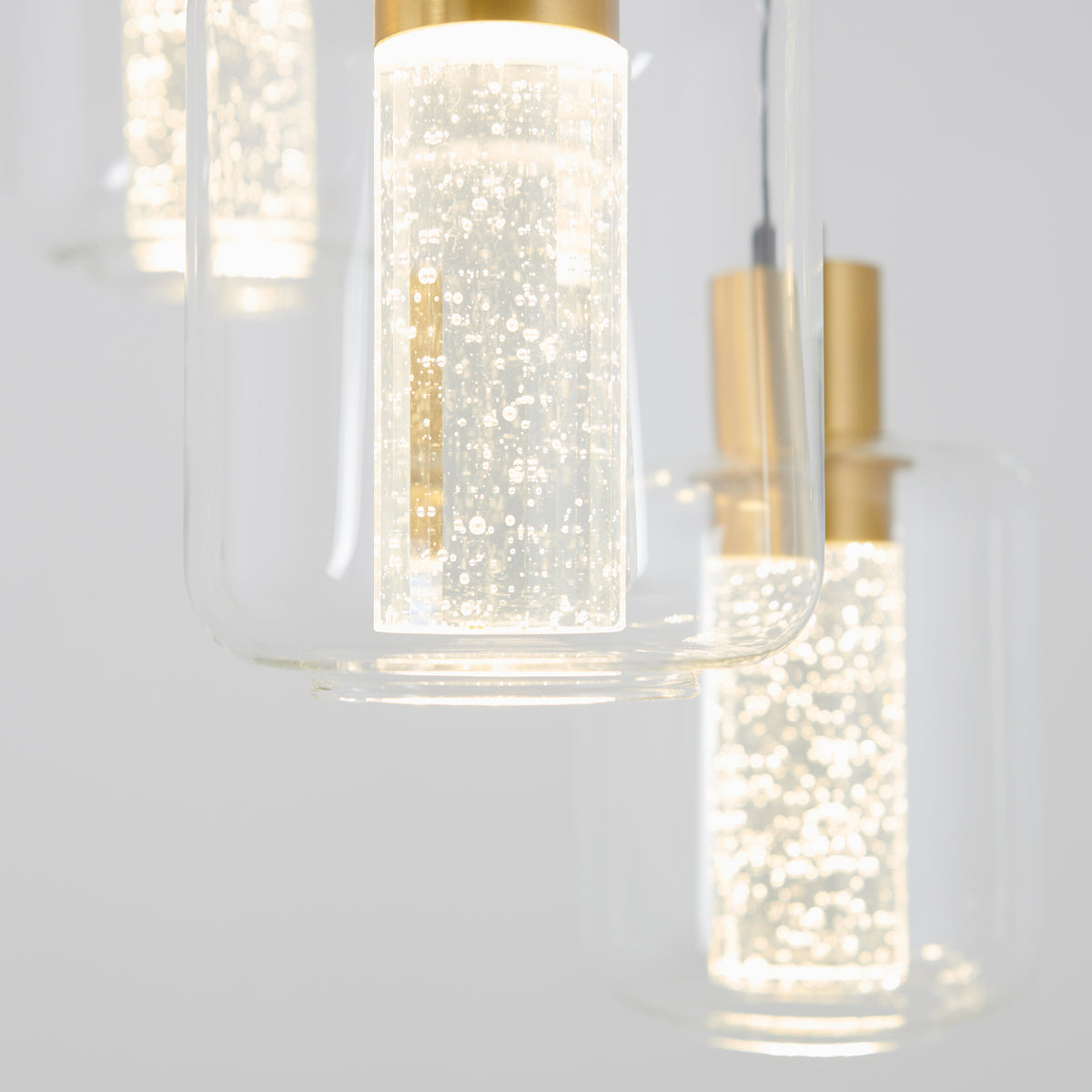 Close-up of the Artika Champagne Globe 4-light LED pendant, featuring illuminated cylindrical glass shades with gold accents and decorative air bubbles, set against a light background.
