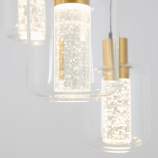 Close-up of the Artika Champagne Globe 4-light LED pendant, featuring illuminated cylindrical glass shades with gold accents and decorative air bubbles, set against a light background.
