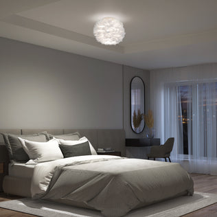 Modern bedroom featuring Artika’s Plume LED flush mount light, a large bed, neutral décor, desk and chair, oval mirror, and floor-to-ceiling windows with sheer curtains framing a dusk sky.