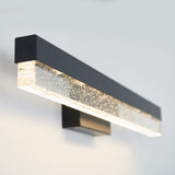 A modern wall light fixture with a black metal frame and a clear, textured glass bar filled with small bubbles, emitting a soft glow against a light-colored wall.