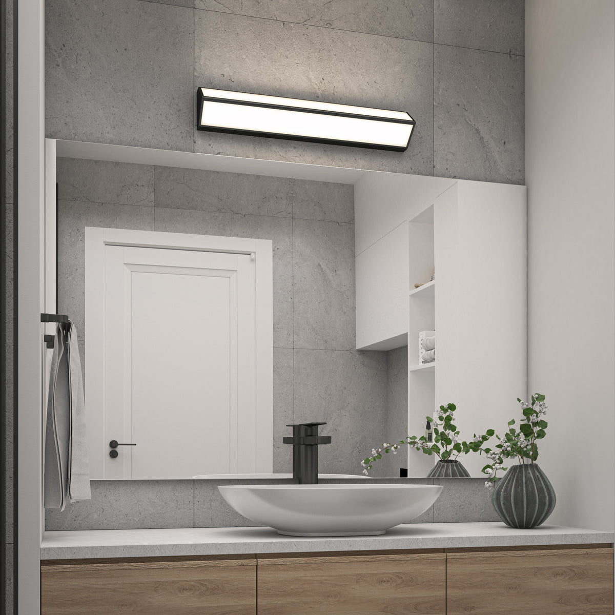 Modern bathroom with a round white sink, black faucet, wooden vanity, and potted plants. A large mirror reflects a white door, and a rectangular light fixture is mounted above the mirror on a gray tiled wall.