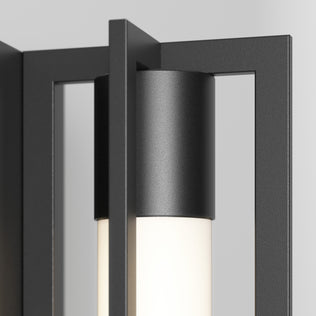 Close-up of the Artika Walt indoor-outdoor LED wall light, featuring a black metal frame and cylindrical frosted glass shade, emitting soft white light against a light gray background.