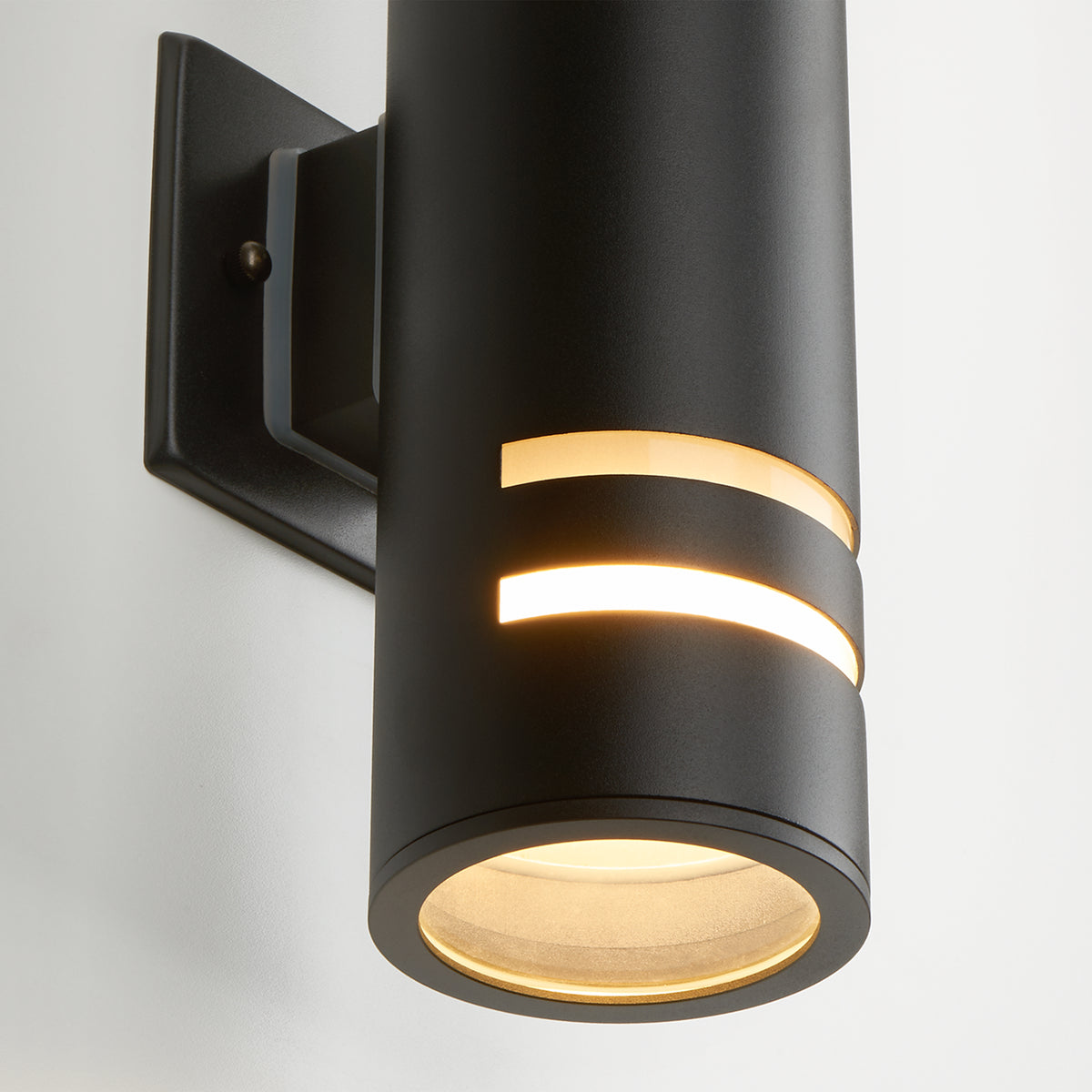 Artika’s V3 indoor-outdoor wall light is a modern black cylindrical sconce with horizontal cutouts that emits warm light, mounted on a square black plate and shown against a white background.