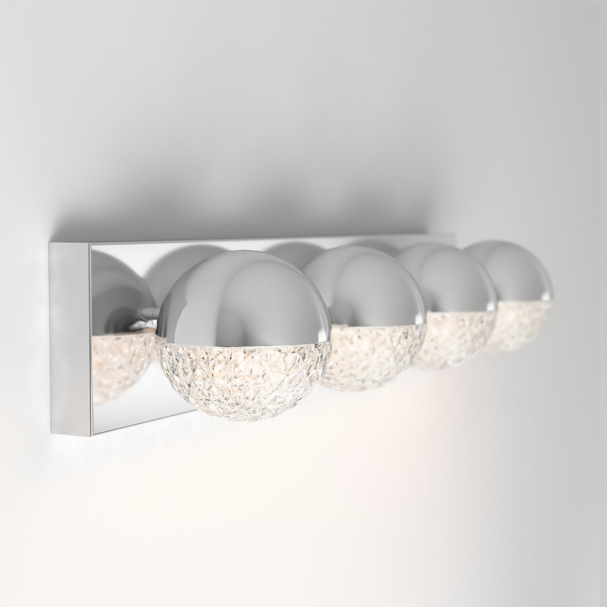 A modern wall-mounted light fixture with four spherical lamps, each half chrome and half textured glass, attached to a shiny rectangular chrome base against a white wall.