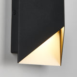 A modern black wall sconce with a geometric design emits a warm light from its angled, partially open bottom, mounted on a white wall.
