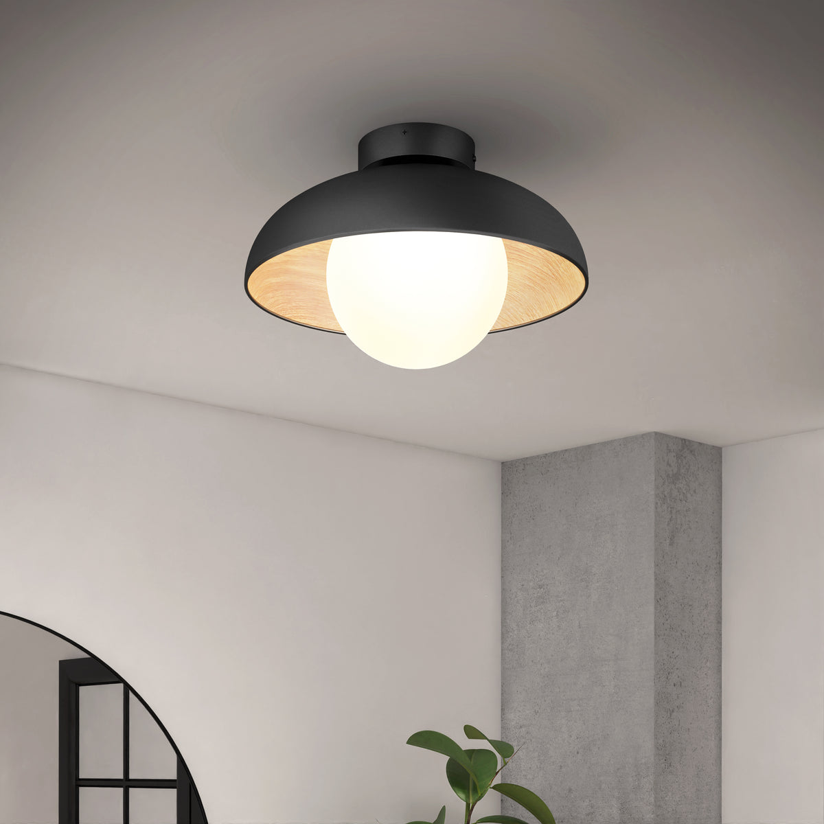 A modern, black, dome-shaped ceiling light with a bright white bulb glows in a minimalist room with gray walls and a small plant visible below.