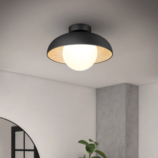 The Artika Boden LED flush mount light 5 CCT, a modern black ceiling fixture with a round white bulb, is mounted on a white ceiling above a gray concrete column, a leafy plant, and an arched window.