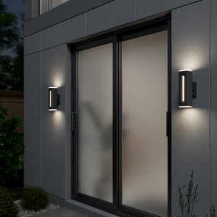 Two Artika Linea Square LED indoor-outdoor wall lights (3 CCT) are mounted on each side of a glass sliding door with frosted panels on a gray exterior, surrounded by lush greenery.