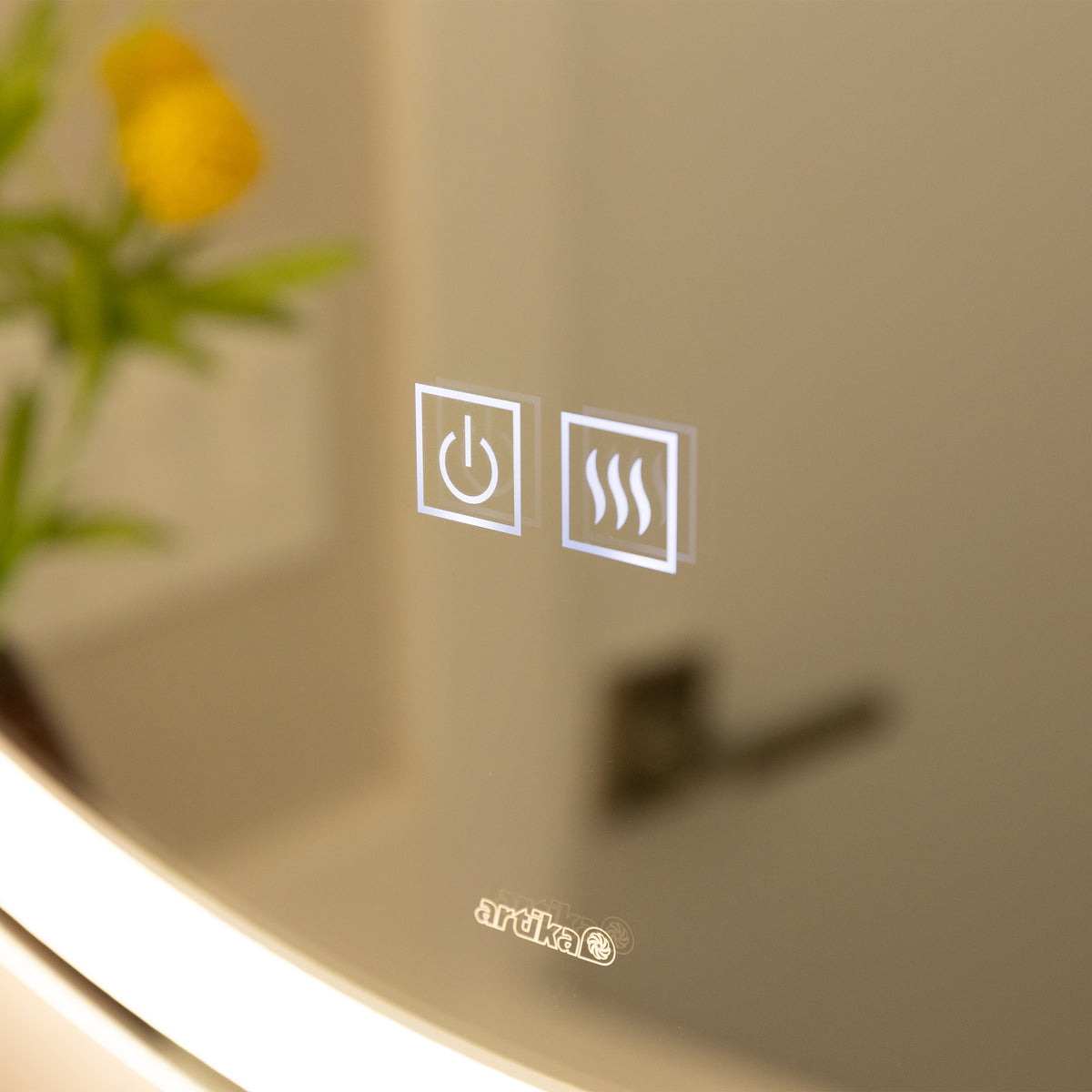Close-up of a mirror with two illuminated touch buttons: a power symbol and a heating/defogger symbol. A blurred yellow flower and green leaves are visible in the reflection. The brand name ardika appears below the buttons.