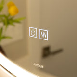 Close-up of a mirror with two illuminated touch buttons: a power symbol and a heating/defogger symbol. A blurred yellow flower and green leaves are visible in the reflection. The brand name ardika appears below the buttons.