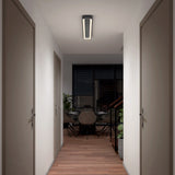 A hallway with wooden flooring and closed doors on both sides leads to a dining area with a round table, chairs, indoor plants, and large windows with dark blinds. A modern ceiling light illuminates the space.