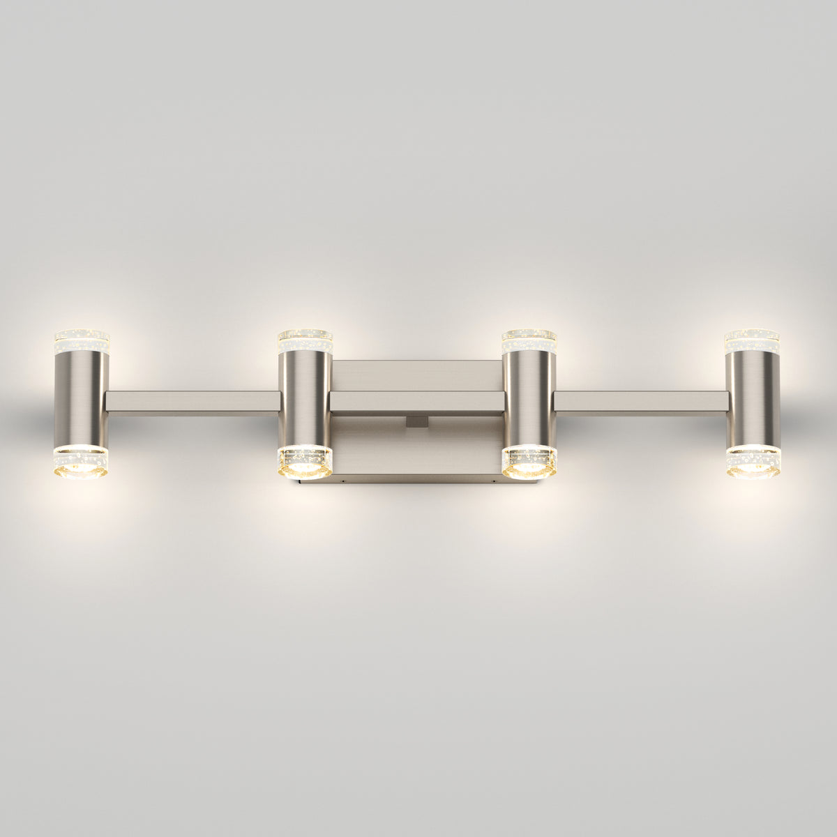 A modern brushed nickel wall sconce with four cylindrical lights arranged horizontally, each with a small round glass accent on the ends, illuminating a light gray wall.