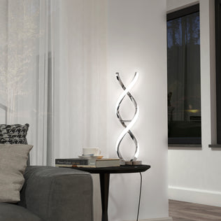 The Artika Swirl LED table lamp with a chrome finish sits on a small black side table beside a gray sofa, accompanied by books and a cup. A large window with sheer white curtains is in the background.