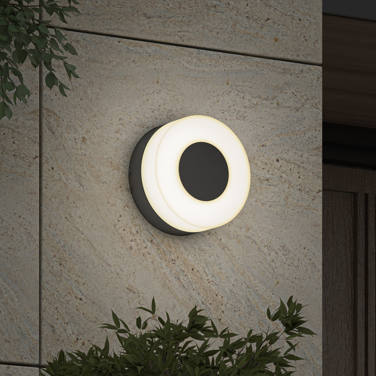 The Artika Ludwig modern LED indoor-outdoor wall light 5CCT, featuring a glowing white ring, is mounted on a textured stone exterior wall and surrounded by green leafy plants.
