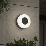 A modern round wall light with a glowing white halo is mounted on a beige stone exterior wall, surrounded by green leafy plants at the edges of the image.