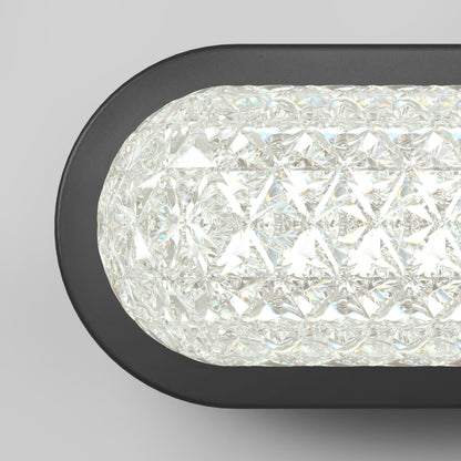 A close-up of the Artika Carat LED contemporary bathroom vanity light 3 CCT, featuring a black frame and textured crystal-like glass, emits bright white light against a plain gray background.