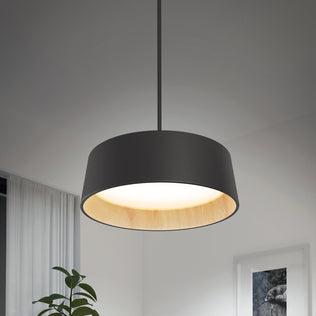 The Alton modern LED pendant light 5 CCT by Artika hangs in a minimalistic room with a leafy green plant, curtain, and framed artwork softly lit in the background. Its black finish features a light wood interior for added style.