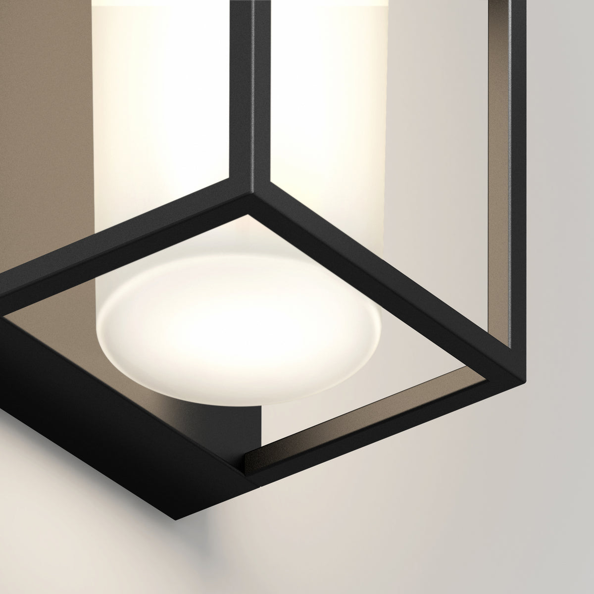 Close-up of a modern wall-mounted light fixture with a black metal frame and a frosted cylindrical glass shade, emitting a soft, warm glow against a light-colored wall.