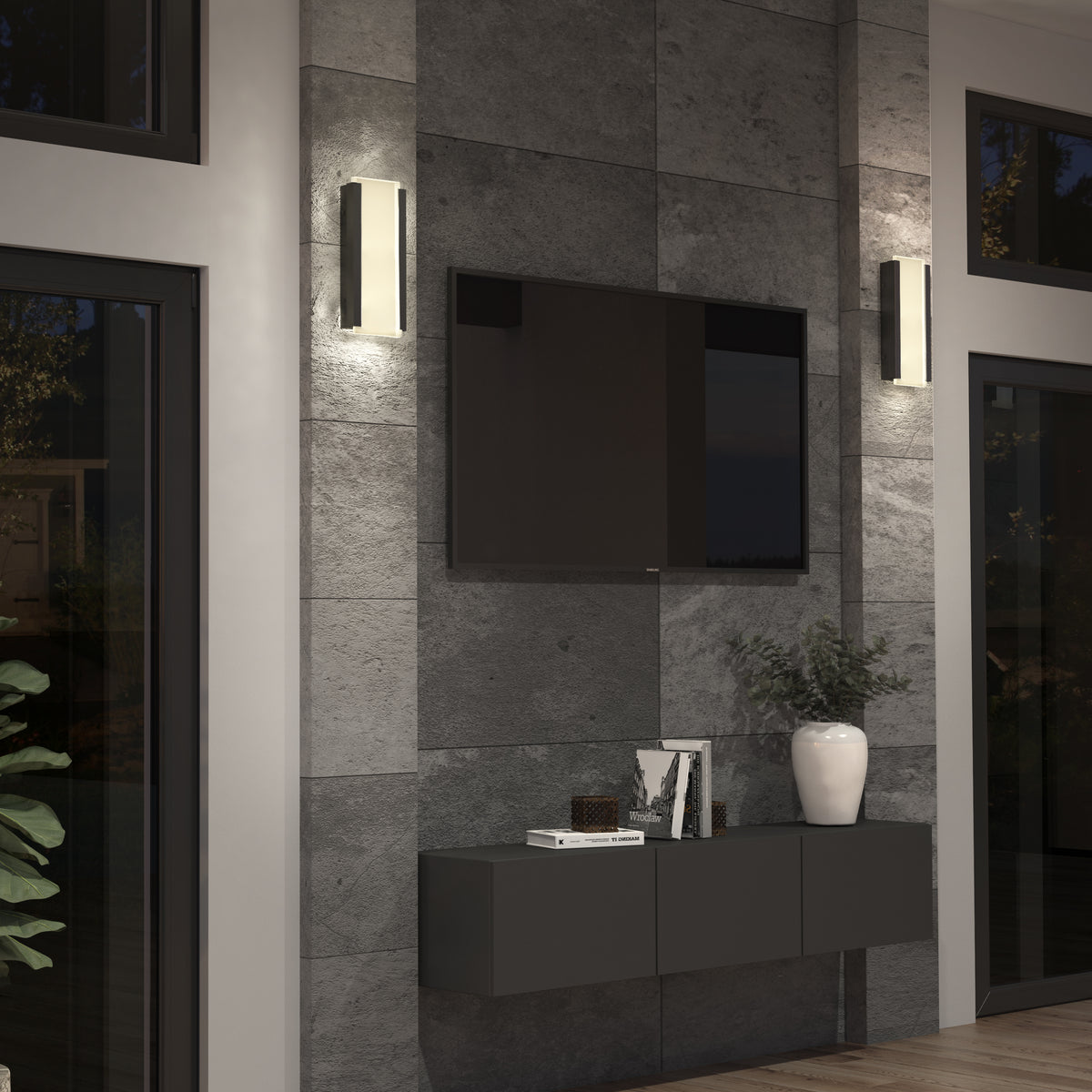 A modern living room with gray stone walls, a wall-mounted TV, two vertical wall lights, a floating black cabinet, a white vase with greenery, and a few books and decor items on the cabinet.