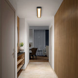 A modern hallway with wooden floors, white walls, and vertical wood paneling on the right. A console with decor is on the left. The hallway leads to a room with a table, chairs, and a large window with curtains.
