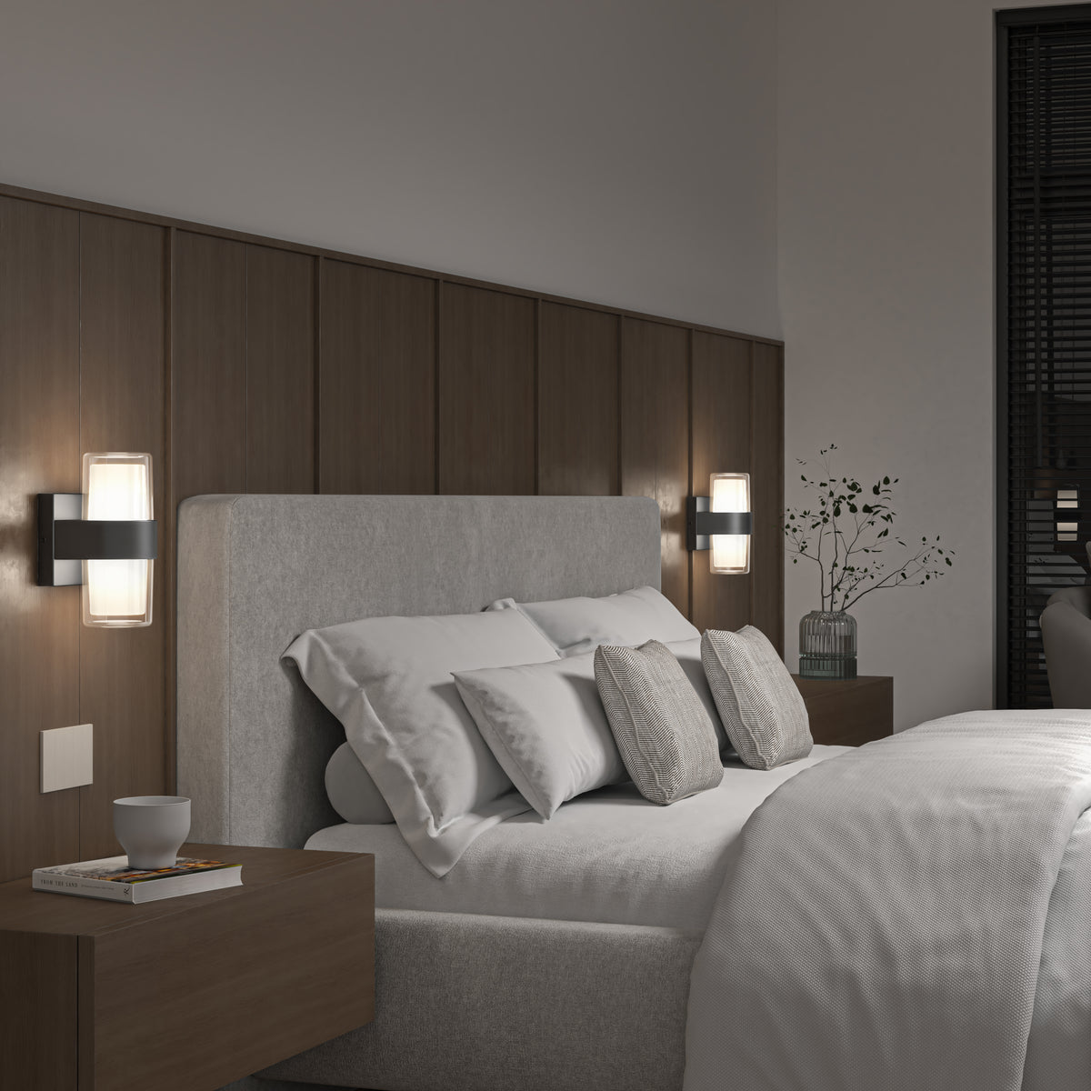 A modern bedroom with a gray upholstered bed, white and gray bedding, two wooden nightstands, wall-mounted lamps, and a vase with branches on one nightstand. The room has wooden paneling and dark window blinds.