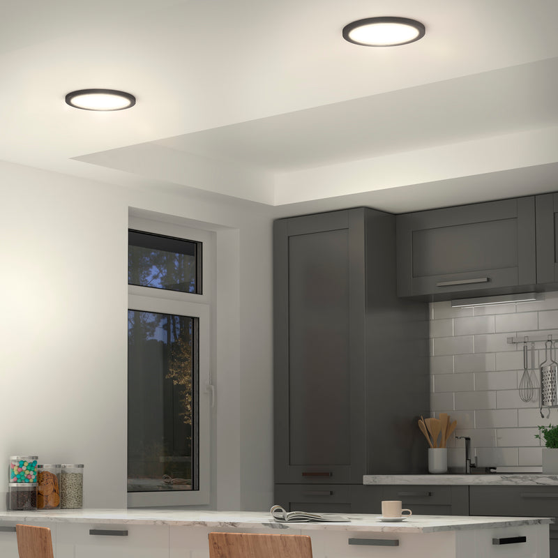 A modern kitchen features Artikas Europa Disk 9 in. LED flush mount lights, gray cabinets, a window, white subway tile backsplash, and a white countertop with a mug and plate. Wooden chairs are partially visible in the foreground.