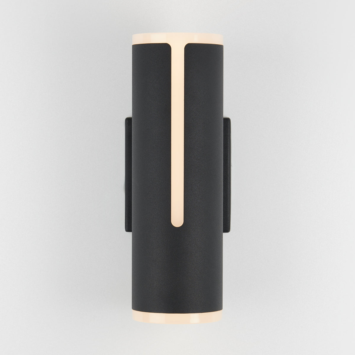 The Artika Linea modern indoor-outdoor LED wall light is a sleek black cylindrical sconce, emitting soft warm light from the top, bottom, and a front vertical strip, shown mounted on a light gray wall.